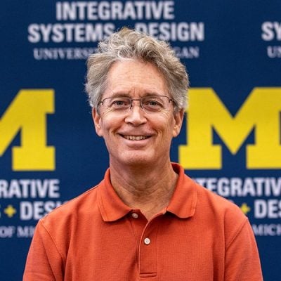 Man with orange dress shirt against U-M logo backdrop. 