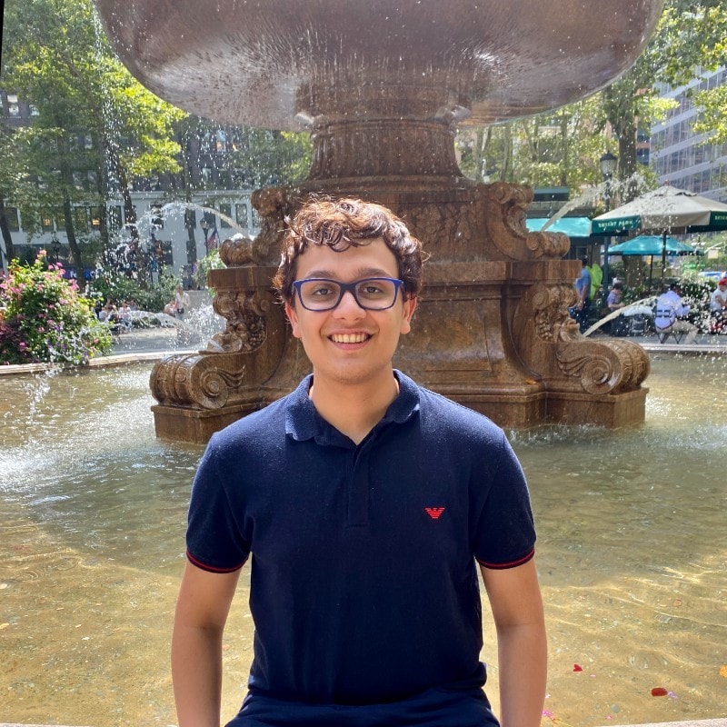 Young man with glasses in blue polo in front of a fountain. 