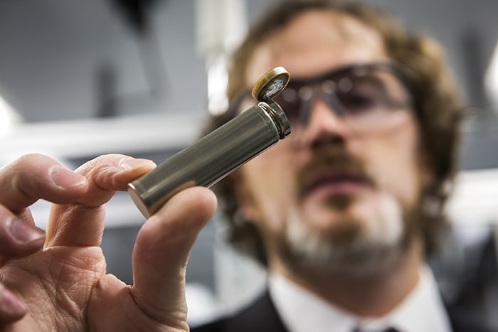 Hand holding cylindrical battery in focus in foreground, with the man in safety goggles out of focus in the background. 